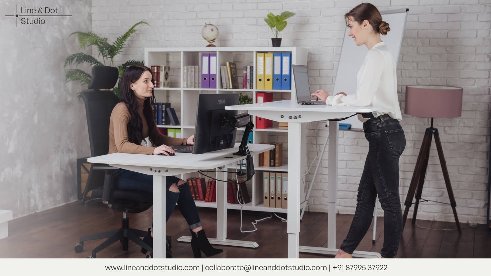 one woman sitting on an ergonomic chair and another woman working on an ergonomic standing desk with shelves around the room