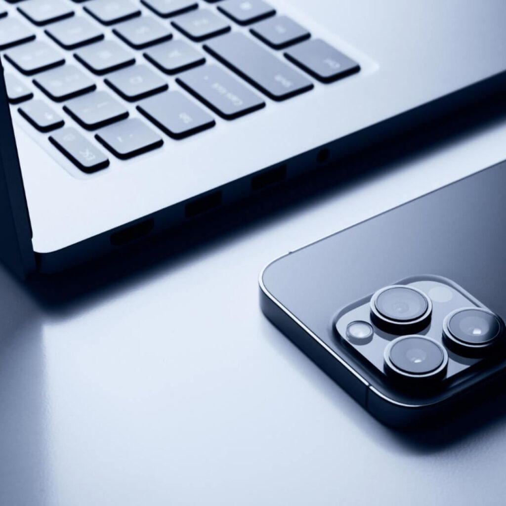 Open Apple MacBook next to iPhone with rear cameras visible, showcasing Apple design aesthetics and product strategy