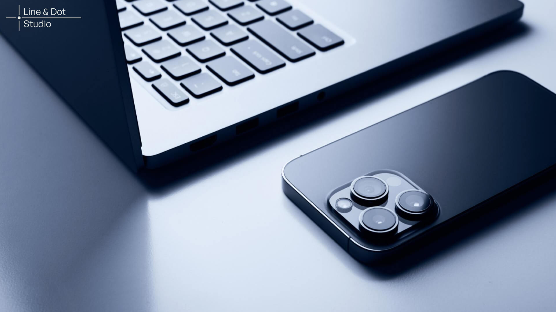 Open Apple MacBook next to iPhone with rear cameras visible, showcasing Apple design aesthetics and product strategy