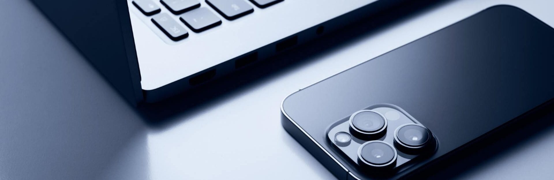 Open Apple MacBook next to iPhone with rear cameras visible, showcasing Apple design aesthetics and product strategy