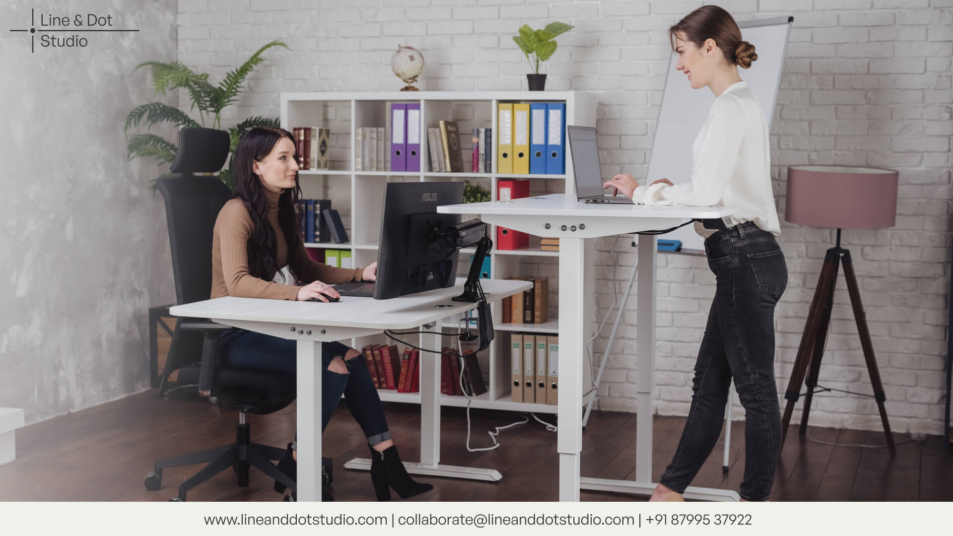 one woman sitting on an ergonomic chair and another woman working on an ergonomic standing desk with shelves around the room